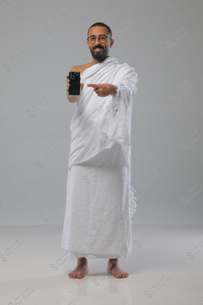 A man wearing traditional white Ihram clothing is standing on a white floor, displaying a smartphone with one hand while pointing at it with the other. He appears happy and is wearing glasses. The background is gray, highlighting the focus on the person and the clothing.