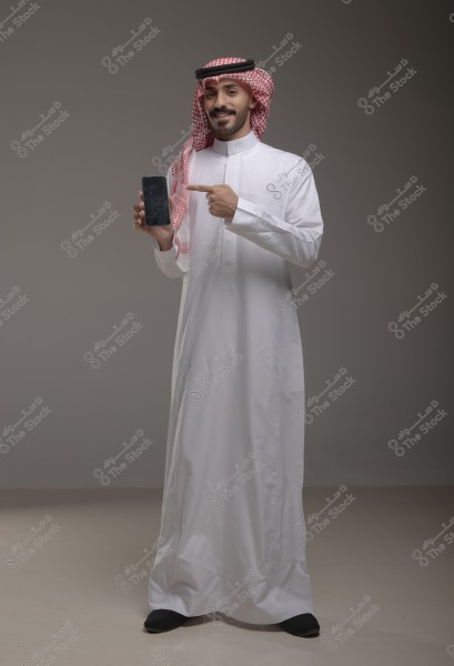 صورة لرجل يرتدي الثوب الأبيض التقليدي والغترة والعقال الخليجيين, يشير بيده إلى هاتف ذكي يحمله في يده الأخرى. يعرض الثوب طابعاً من الأزياء التقليدية الشائعة في المملكة العربية السعودية.