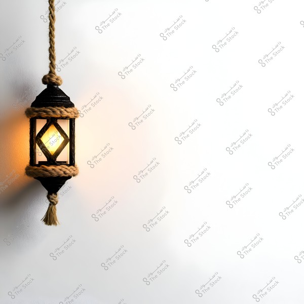 Image of a traditional lantern hanging by a thick rope, emitting a warm glow against a white wall. The design of the lantern and the rope embellishments reflect a traditional Middle Eastern style.