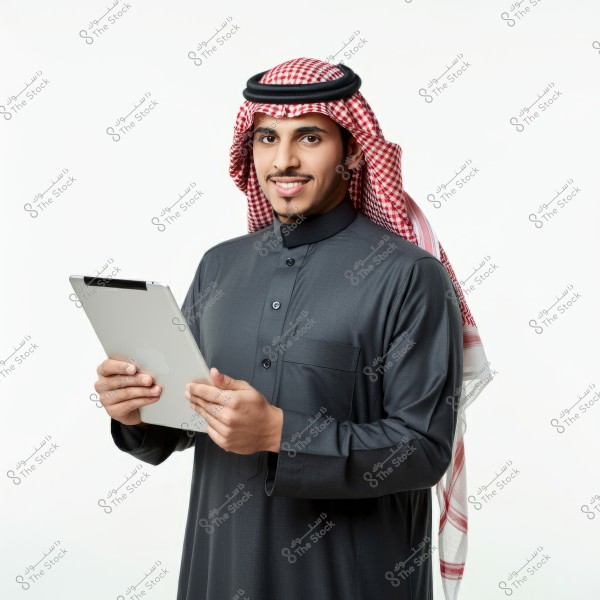 A portrait of a man wearing a traditional Saudi thobe and a red and white checkered headscarf, standing and smiling while holding a tablet. The attire suggests a traditional Saudi appearance.