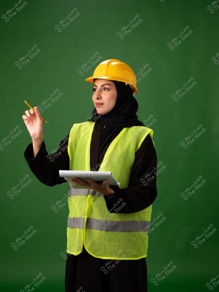 Image of a woman wearing engineering work attire, including a yellow safety vest and a yellow hard hat over a black hijab. She is holding a pen and a notebook, standing against a green background and appears focused.