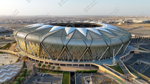 An aerial view of a modern stadium with a distinctive architectural design, featuring a large metallic roof interwoven with geometric shapes and patterns. It is surrounded by open areas, paved roads, and scattered cars. In the background, buildings and vast land areas suggest it is located in a large urban area.