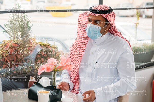 رجل سعودي يرتدي الزي السعودي التقليدي و الكمامة يجلس في المقهى يتحدث لأحدهم وتظهر على الطاولة أمامه مزهرية وعلبة مناديل وكوب قهوة وإلى يمينه زجاج النافذة وتظهر خلفها مجموعة من النباتات