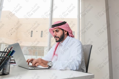 رجل سعودي يرتدي الزي السعودي التقليدي يعمل في مكتب بواجهة زجاجية ويستخدم قلم رصاص لتدوين الملاحظات