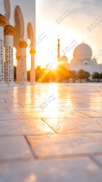 The image shows a large mosque with white domes and decorative columns, with the sunset in the background casting golden shadows on the shiny marble floors. A tall minaret is visible among the distinctive Islamic architecture and green trees in the background.