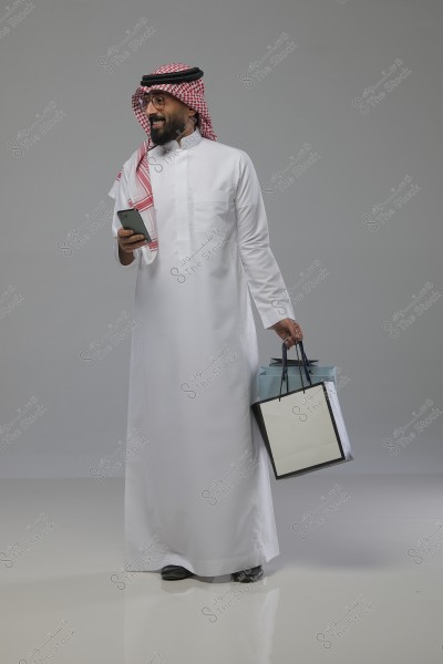 The image shows a man in traditional Saudi attire, consisting of a white thobe and a red and white shemagh. He is holding a mobile phone in his right hand and two shopping bags in his other hand. The man is smiling and looking away, with a simple gray background.