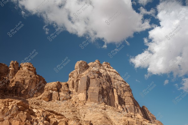 A natural landscape featuring massive rocky mountains under a clear blue sky with scattered white clouds. The rocks exhibit various shades of brown, highlighting the grandeur and geological details of nature.