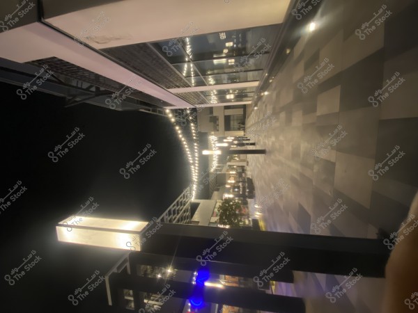 A flat, illuminated pathway at night with modern light posts. The ground is tiled with various geometric patterns. Modern buildings with large windows are on either side, reflecting a contemporary design aesthetic.\r\n\r\n###