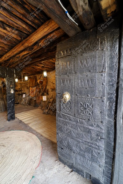 The image shows the entrance of a traditional house with a wooden door intricately carved with geometric and brass designs. Inside, the ceiling is supported by wooden beams, and the floor is covered with mats, illuminated by hanging lanterns. There are old tools mounted on the walls inside the house.