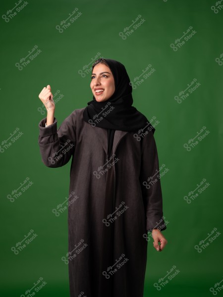 A portrait of a woman wearing a black abaya and a hijab, standing and smiling with her hand raised as if expressing victory or joy. The background is green, providing a clear contrast for the image.