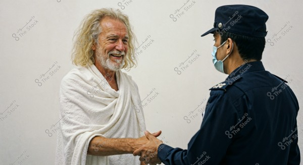 The image shows two men shaking hands. The first man is wearing a white ihram, while the second man is in a blue uniform with a face mask. The background is plain white.
