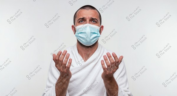 Portrait of a person wearing a blue medical mask and a white robe, raising their hands upwards against a white background. The person appears to be in a state of focus or confusion.
