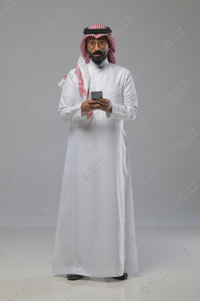 A portrait of a man wearing traditional Saudi attire, consisting of a white thobe and a red and white keffiyeh with a black agal. The man is standing, holding a smartphone in his hands, and looking at it. The background is a simple gray.