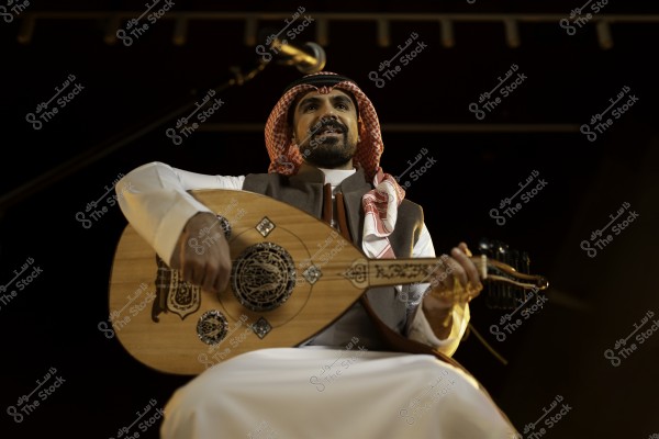 A oud player sitting on stage wearing traditional Gulf attire, playing the oud under a microphone.