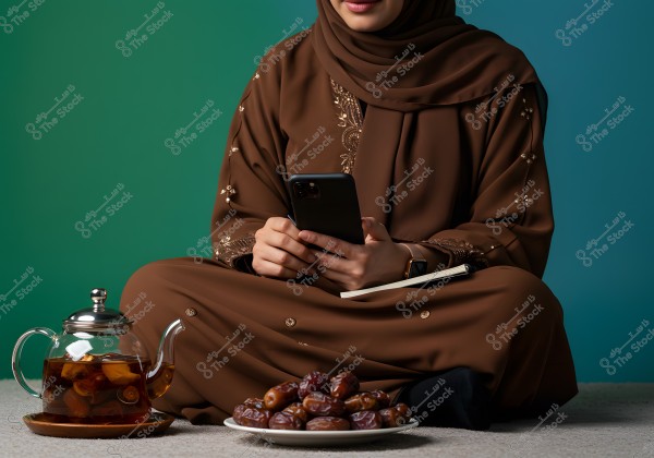 A woman wearing a brown abaya with golden embroidery, sitting on the floor while holding a mobile phone. In front of her on the floor, there\'s a glass teapot with tea, and a plate of dates. The background gradients from green to blue, adding a modern touch to the image.