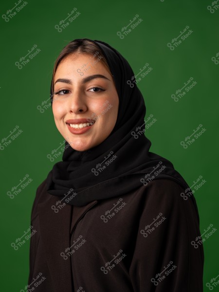 صورة لامرأة ترتدي حجابًا أسود وتبتسم, مع خلفية خضراء.
-