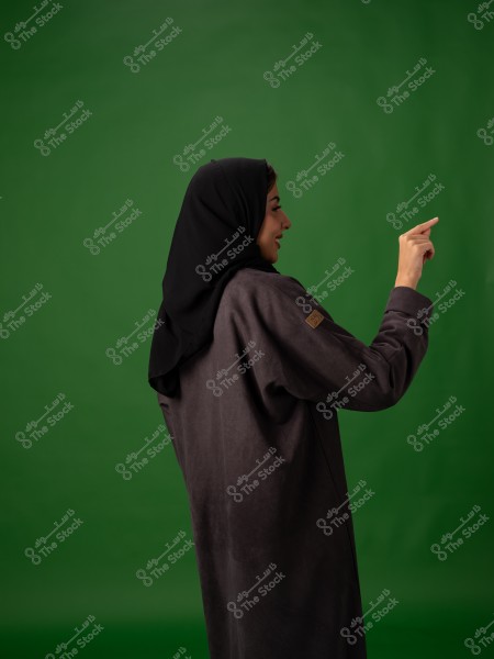 امرأة ترتدي حجابًا وعباءة بنية تقف وتشير بإصبعها على خلفية خضراء.