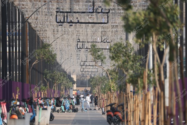 يظهر في الصورة شارع مزين بأضواء معلقة بشكل جاذب, تحيط به أشجار مصطفة على الجانبين. تتواجد العديد من الدراجات الكهربائية على الرصيف, وبعض الأشخاص يسيرون في الخلفية, يرتدي أحدهم ثوبًا أبيض تقليديًا. الأجواء توحي بمدينة حديثة في منطقة الشرق الأوسط.