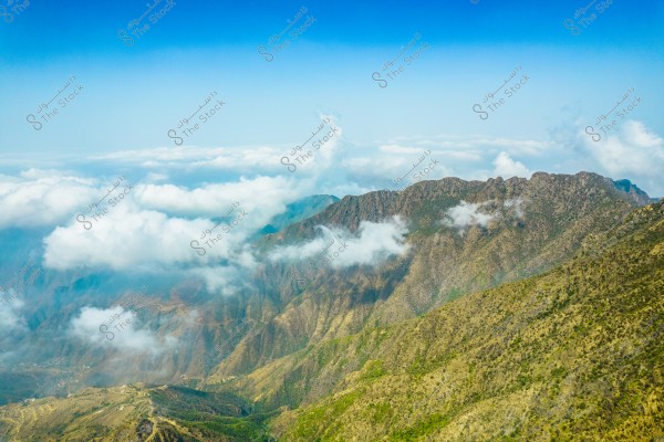 A natural landscape featuring a rugged mountain range covered with vegetation. White clouds partially obscure the mountains, with a clear blue sky in the background. The mountains display varying shades of green and brown, highlighting the area\'s biodiversity.