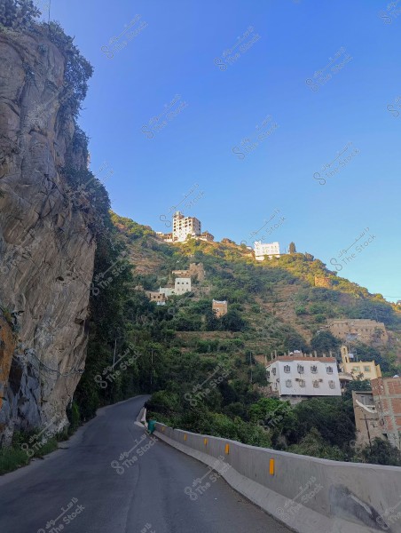 A narrow mountain road with houses built on a hillside under a clear blue sky. Buildings are scattered along the plant-covered slope.