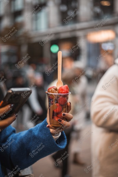 A person holding a plastic cup filled with strawberries and chocolate sauce, with a wooden spoon on top. The background shows a busy street.