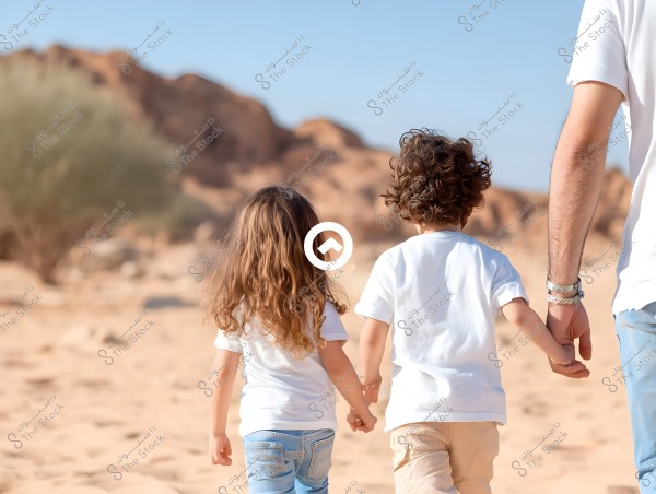 The image shows three people from behind walking in a desert area. Two children wearing white T-shirts hold hands with a man who resembles a father, wearing a white T-shirt and jeans. The background features sandy hills and a clear blue sky.