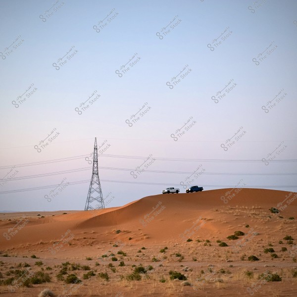 A scene of a sandy desert dotted with small plants, featuring a high sand dune with two cars driving on it. In the background, there\'s a large power tower and clear blue skies.
