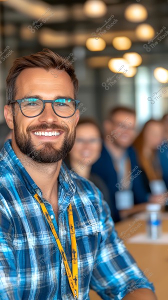 Image of a man wearing glasses and a blue checkered shirt, smiling. In the background, there are blurred people sitting in an office setting with round ceiling lights. The man has a yellow ID lanyard around his neck.