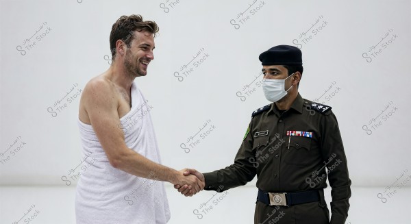A man wearing a white Ihram garment shakes hands with another man in a green military uniform with a cap and face mask, adorned with badges and ribbons, against a plain white background.