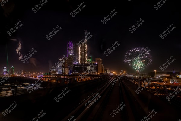 The image shows a nighttime scene of a city sparkling with lights and tall buildings. In the sky, colorful fireworks burst near the buildings. A bridge or highway runs through the scene towards the foreground, adding depth to the image. The buildings are lit with various colors, and some are adorned with patterns and lights that brighten the darkness.