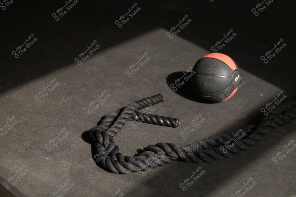 The image shows a black and red medicine ball labeled \"4 KG\" and a rope with black rubber handles, placed on a dark floor partially illuminated by sunlight. The setting conveys a sense of exercise and fitness.
