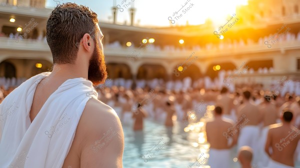 An image of a man wearing a white Ihram standing in the courtyard of the Masjid al-Haram in Mecca, with a crowd of people also in Ihram attire beneath him. The sun is shining in the background, creating a warm and bright scene.