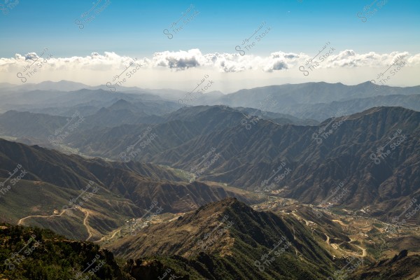 An image of the Al Soudah mountain in Saudi Arabia, featuring a range of green mountains stretching to the horizon with scattered cloud formations in the blue sky. Roads wind around the mountains, clearly depicting the terrain of the area.