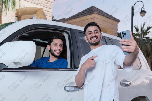 A snapshot of two young Saudis chatting and having fun on the road, their features showing joy and happiness, taking a selfie with a mobile phone