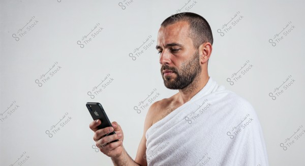 A portrait of a man wearing a white Ihram, holding a smartphone in his hand and looking at it. The background is white, and the face is shown from the right side. The Ihram suggests that the photo might have been taken before performing Hajj or Umrah.