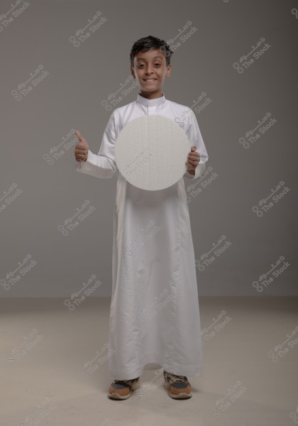 A portrait of a boy wearing a white thobe, holding a white circle with both hands, while giving a thumbs-up with his other hand. He is smiling, set against a neutral gray backdrop. The boy is wearing sports shoes and is standing upright.