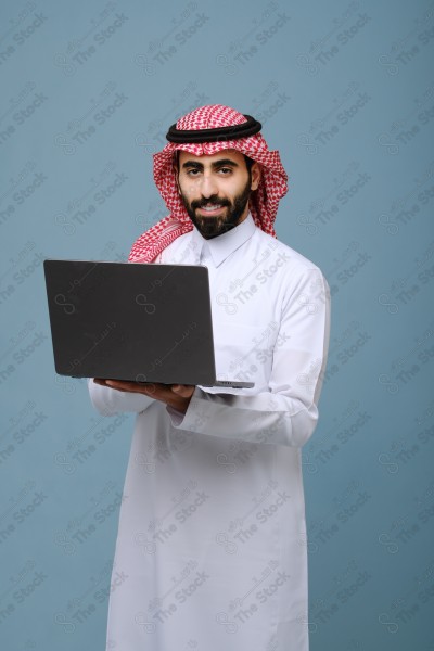 بورتريه شاب سعودي يرتدي الزي السعودي ينظر إلى جهاز المحمول وتظهر على ملامحة السعادة ، يقف على خلفية سماوية