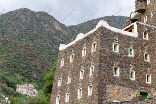 An ancient building built of stones, in the middle is a group of windows surrounded by white paint, behind which mountains are covered with a group of trees،Rijal Almaa Heritage Village in the Asir region