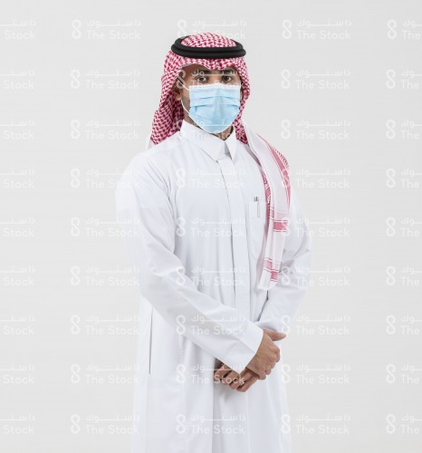 شاب سعودي يرتدي الزي السعودي الرسمي على خلفية بيضاء وهو واضع الكمامة على وجهة.