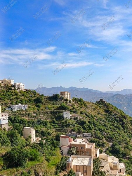 An image of a natural landscape in a mountainous area with terraced fields covered in green vegetation. Traditional buildings are scattered across the mountain, with some in light brown and others in white. In the background, a range of low mountains is visible under a wide blue sky with wispy clouds.