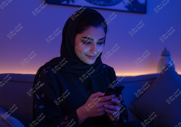 A portrait of a woman sitting on a couch, wearing a black abaya and hijab. She is looking at her mobile phone and smiling in a dimly lit environment with blue and purple lighting. A portion of a cushion is visible beside her, with home decor in the background.