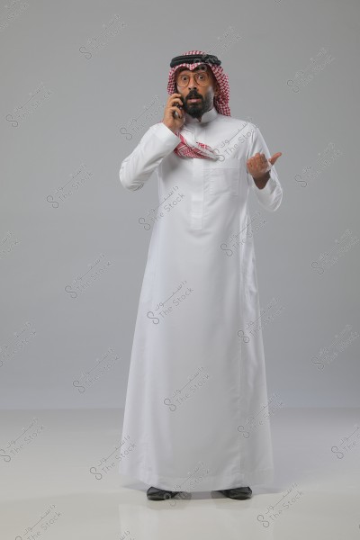 Image of a man wearing a traditional Saudi white thobe with a red checkered shemagh and a black agal. He appears to be talking on a mobile phone with a surprised expression, pointing with his other hand. He stands in a studio with neutral lighting and a gray background.