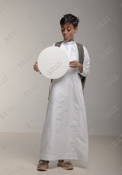 Image of a boy holding a large white circle. The boy is wearing a traditional white thobe and appears to be smiling with curiosity. He has a backpack and sports shoes. The background is smooth and light grey.