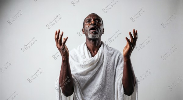 An image of a man of African descent wearing traditional white Ihram clothing typically used for Hajj or Umrah. The person is raising his hands in a prayer-like position, appearing focused and serene. The background is white and neutral.