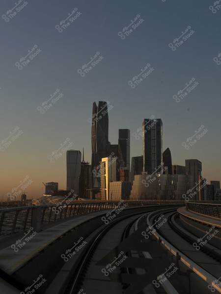 A view of a modern city skyline at sunset, featuring the King Abdullah Financial District towers in Riyadh, Saudi Arabia. The image showcases a cluster of tall buildings with reflective glass, and in the foreground, there is a railway track leading into the city. The sky is blue with a golden hue from the sunset.