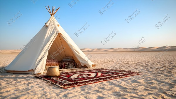 A white tent stands in the middle of a sandy desert under a clear blue sky. In front of the tent is a traditional rug with red and white patterns and a utility basket. The background features vast stretches of dense sand, indicating a wide desert landscape.