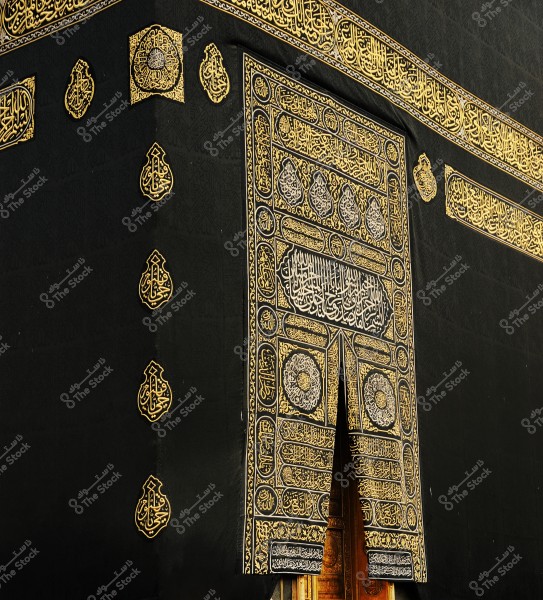 Image of a section of the Kaaba in Mecca, showcasing the black Kiswah adorned with golden Arabic calligraphy. The image highlights the intricate embroidered door with golden designs and script on the black fabric, and the decorative Quranic texts above the Kaaba.
