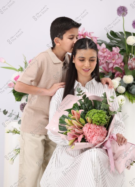 A young boy is kissing a woman\'s head. The woman is wearing a striped dress and holding a bouquet of colorful flowers, including lilies, carnations, and green leaves. In the background, there are various floral arrangements with pink and white flowers against a white background.