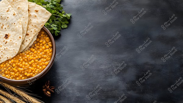 An image of a bowl filled with orange lentil stew topped with four pieces of flatbread. The bowl is placed on a black surface, with some green herbs and wheat stalks beside it.