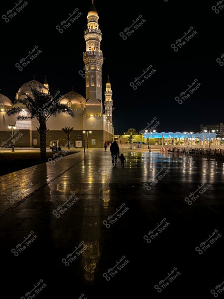 An image depicting the courtyard of a mosque illuminated at night, with lights reflecting off the shiny marble floor. Several minarets and domes of the mosque are visible, along with a palm tree in the foreground. Two people are walking in the courtyard, one of them a small child. The sky is black, and the scene has a serene atmosphere.